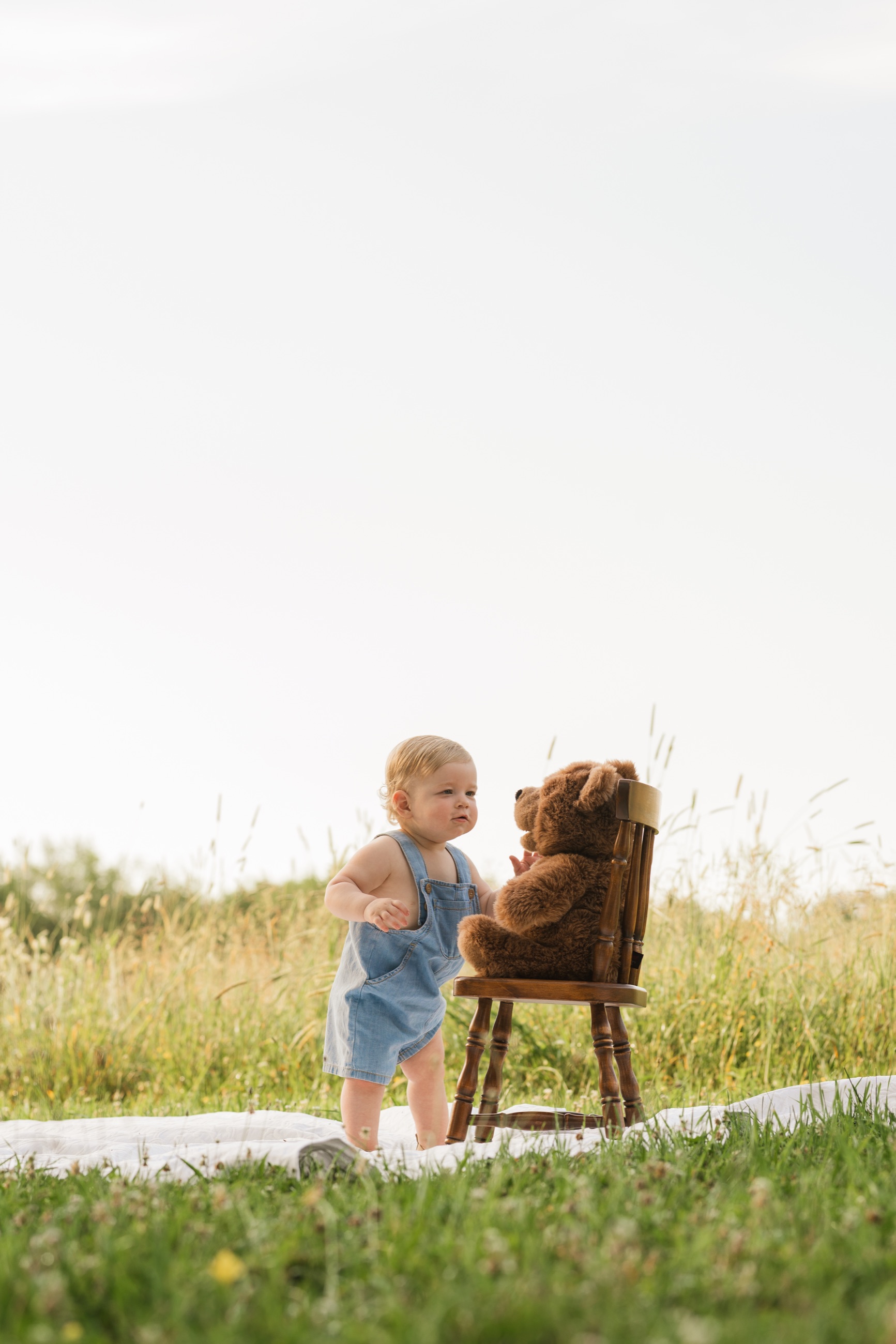 Young child standing beside a teddy bear on a wooden chair in a summer field